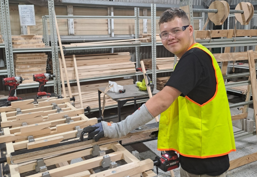Eddie smiling at the camera while holding a woodwork tool and wearing safety goggles at his new workplace.